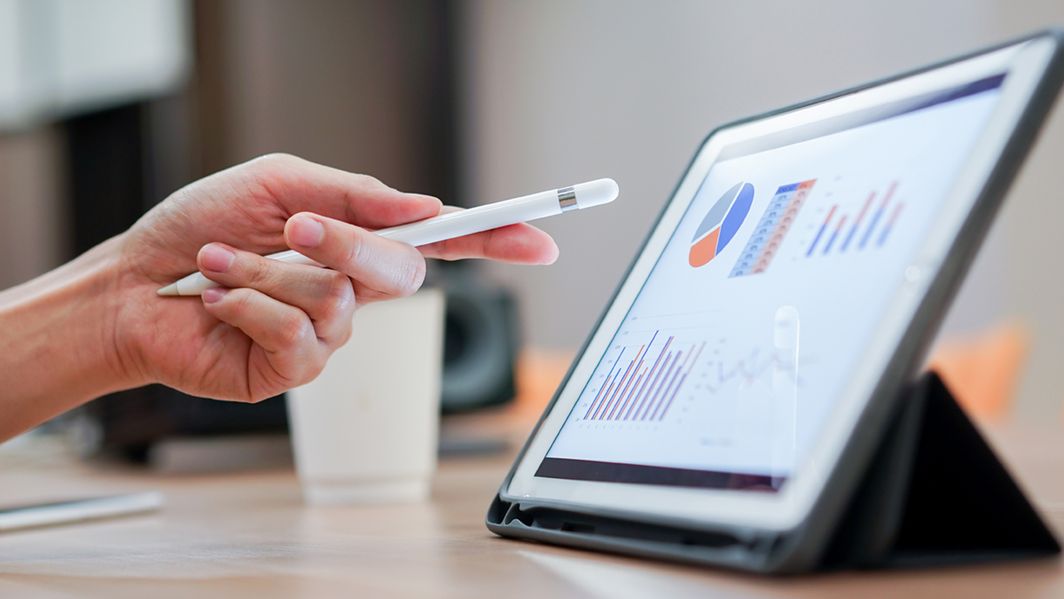 Ein Tablet steht rechts auf dem Tisch. Eine männliche Hand zeigt mit einem Stift auf die Infografiken auf dem Tabletscreen.