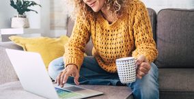 Eine Frau in gelben Pullover sitzt vor einem Laptop mit einer Kaffeetasse in der linken Hand