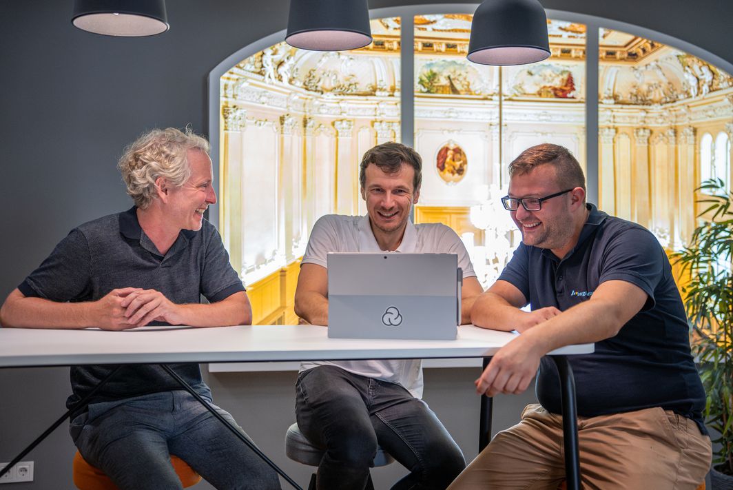 3 Personen sitzen an einem Tisch und schauen gemeinsam auf ein Notebook