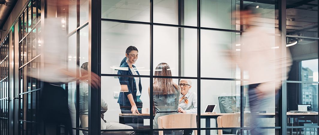 Mehrere Menschen sitzen in einem Büro während andere Mitarbeitende daran vorbeilaufen