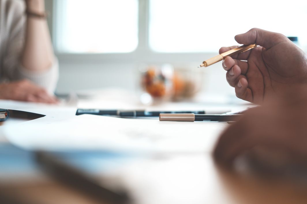 Ein Konferenztisch mit Unterlagen und Stiften. Im Hintergrund ist ein Fenster, rechts sitzt ein Mensch in einem weißen Pullover. Rechts sieht man zwei Hände, eine Hand ist im Fokus und hält einen Stift.